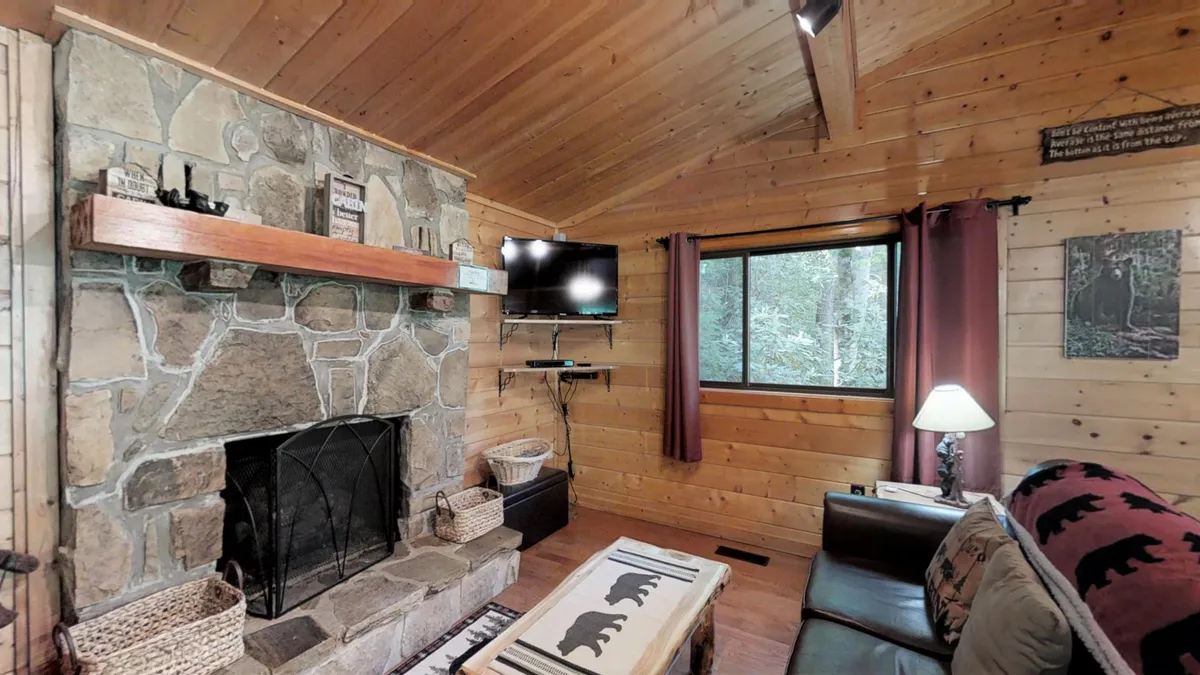 A comfortable living space with a plush sofa, patterned throw, stone fireplace, wood-beamed ceiling, TV, and large window offering views of the surrounding nature. The setting feels cozy and inviting, perfect for relaxing or watching shows together.