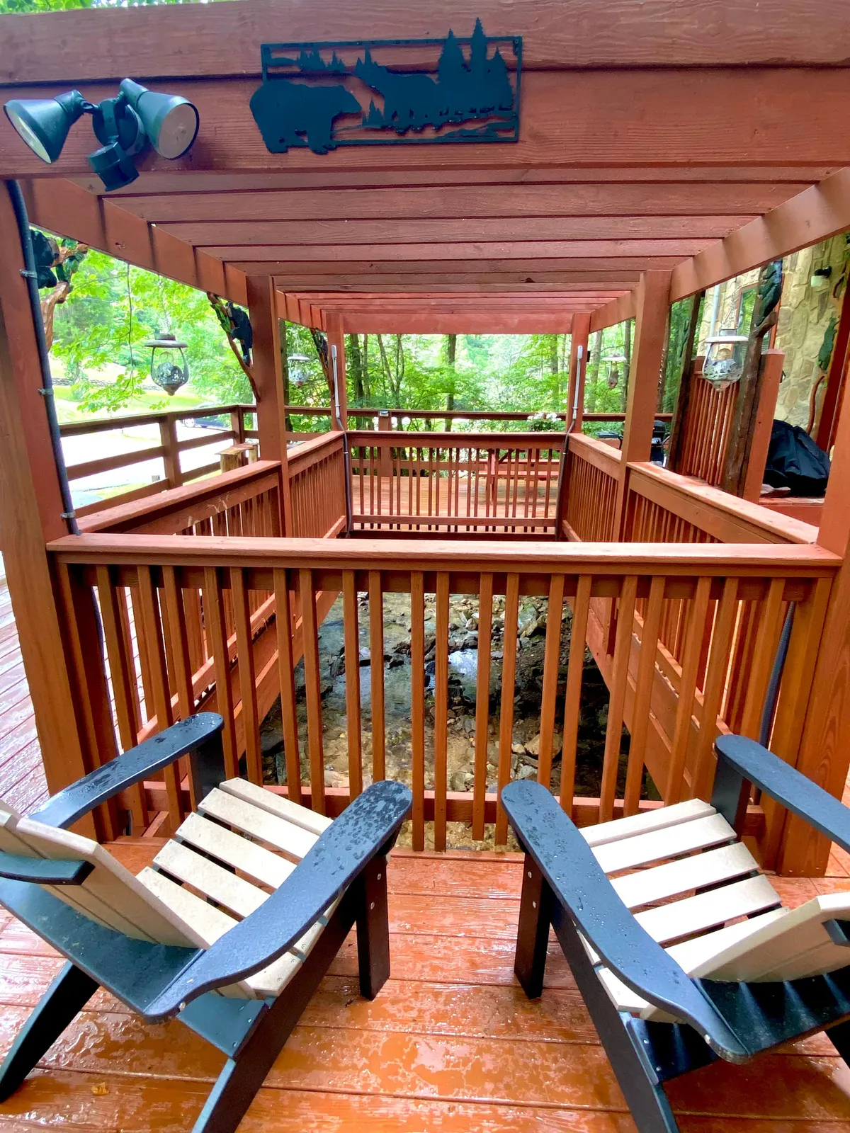 The photo shows a wooden deck perched above a rocky stream, furnished with outdoor chairs and decorative metal art. The scene is enveloped in greenery, conveying an intimate and peaceful environment ideal for couples.