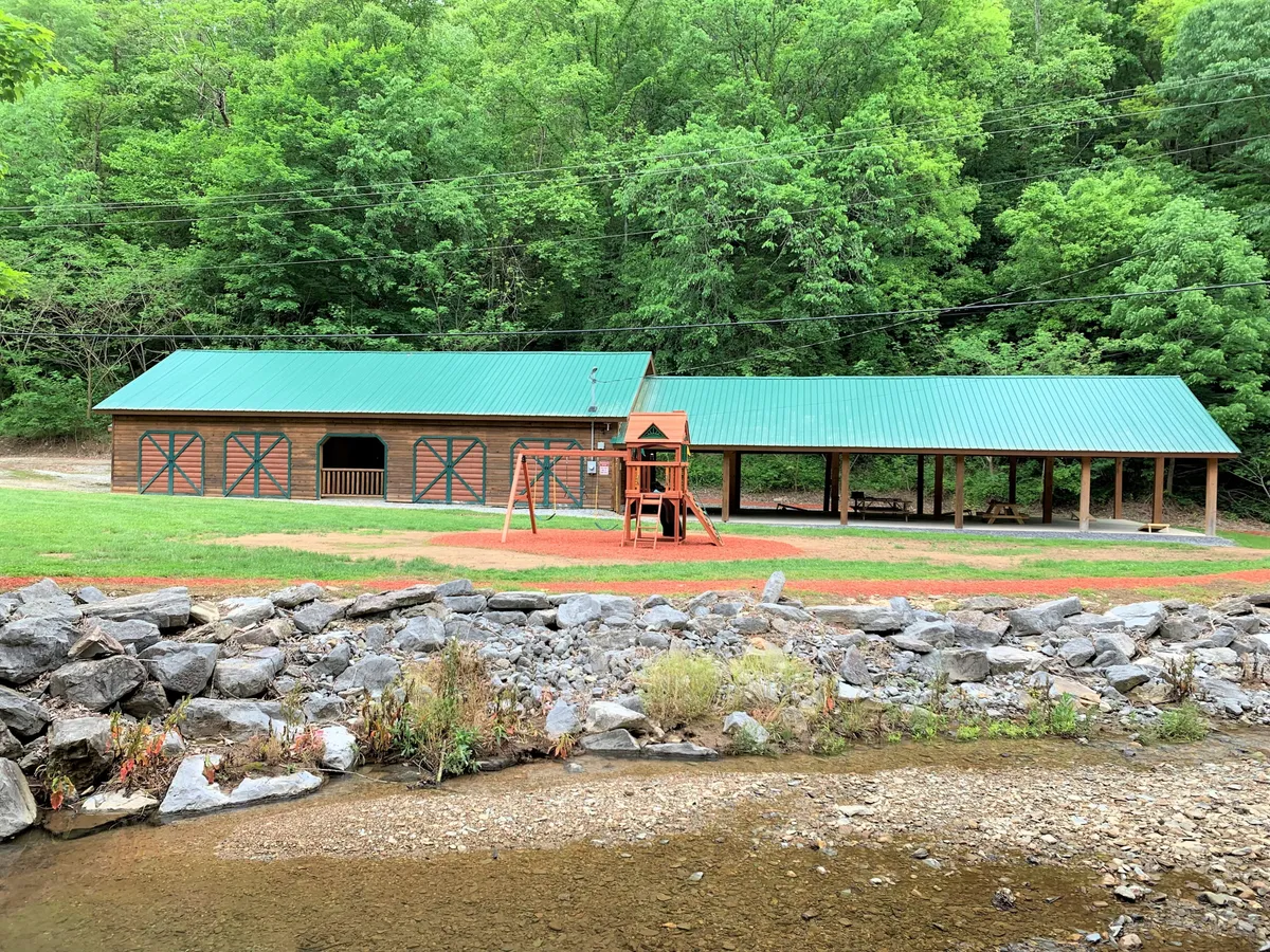 A vibrant outdoor playground set against a rustic wooden building with a green roof, surrounded by lush natural landscaping and a rocky riverbank. Children can be seen playing, while families gather nearby, and the riverbank offers opportunities for peaceful nature exploration.