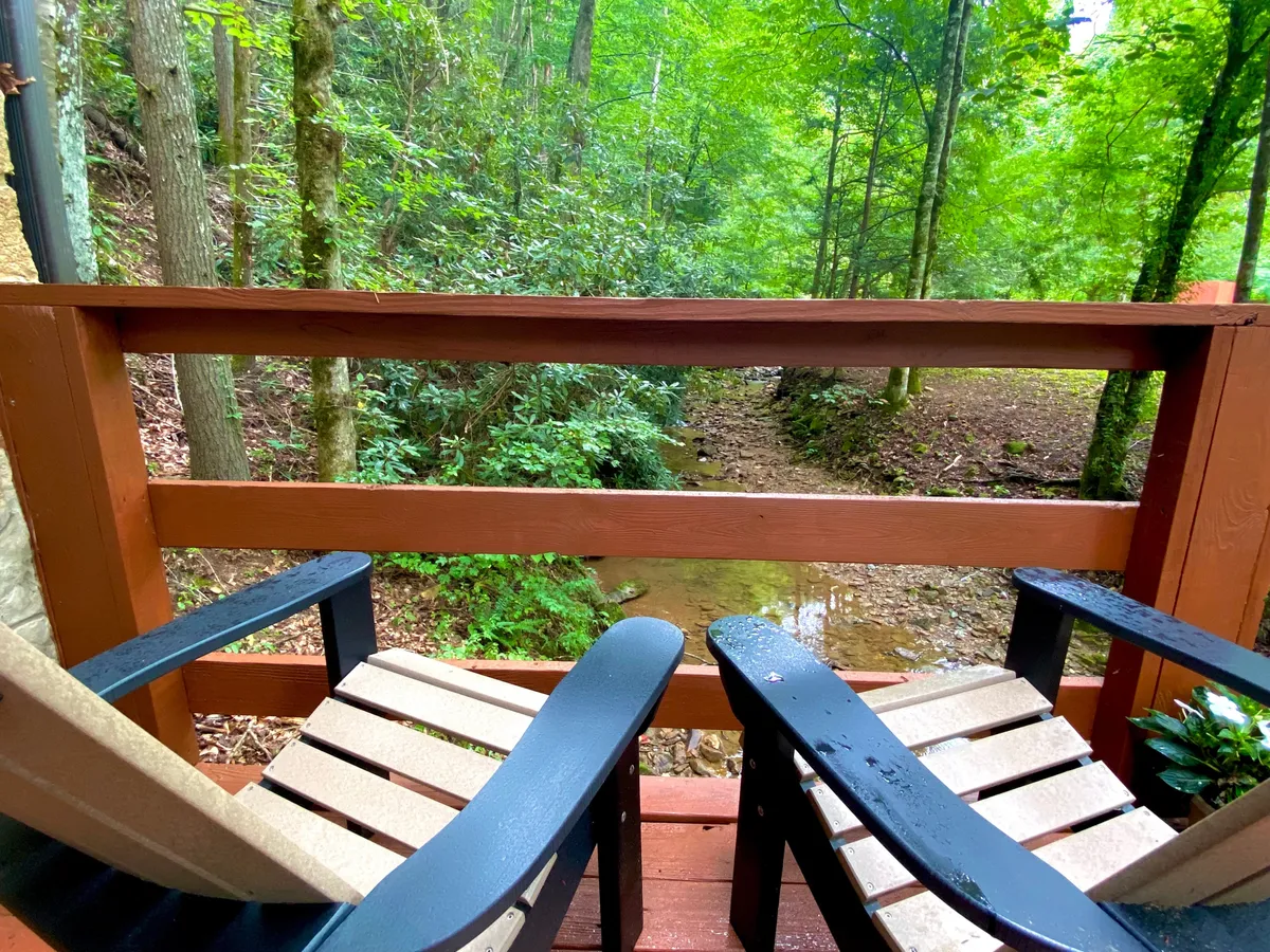 This image captures a wooden porch overlooking a rocky creek, with outdoor chairs positioned for relaxing and taking in the lush greenery. The tranquil natural setting is perfect for enjoying the sounds of the babbling creek and observing local wildlife.