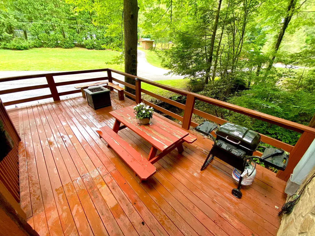 A welcoming wooden deck set amidst lush greenery, featuring a picnic table, grill, and a vibrant flower arrangement. The surrounding trees create a natural, peaceful atmosphere, while the spacious deck invites families to gather and dine outdoors together.