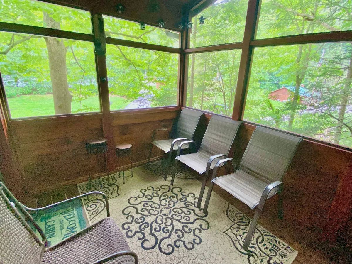 A screened-in porch with comfortable seating, wooden walls, and large screened windows that offer peaceful views of the greenery outside. The relaxed arrangement invites lounging while still immersed in nature.