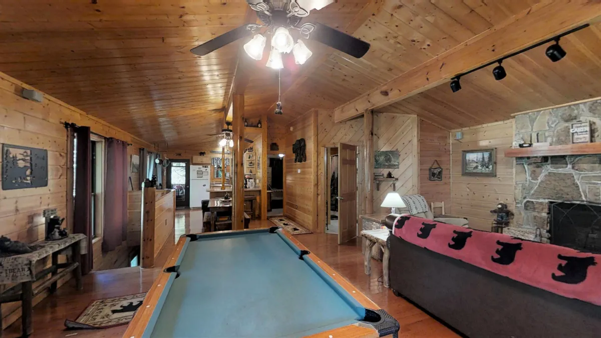 A spacious, open-concept interior featuring a pool table at the center, a striking stone fireplace, wooden ceilings, and a view toward the dining area. The space is set up for both relaxation and lively entertainment, with cozy seating and warm lighting.