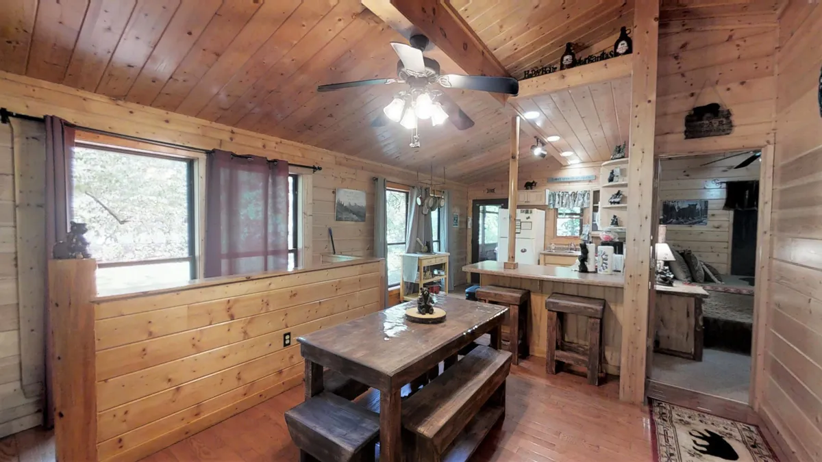 A spacious dining area with a large wooden table, ceiling fan, wood-paneled walls, and expansive windows letting in natural light. The table is set up to host family meals, game nights, or group gatherings in a warm, communal atmosphere.
