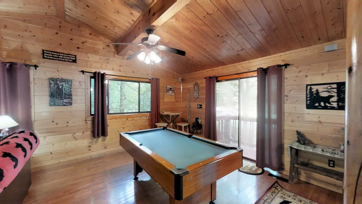 A spacious interior with a pool table, large windows framing woodland views, wood-paneled walls, a ceiling fan, and comfortable seating. The combination of recreation and relaxation elements highlights the cabin's modern amenities.