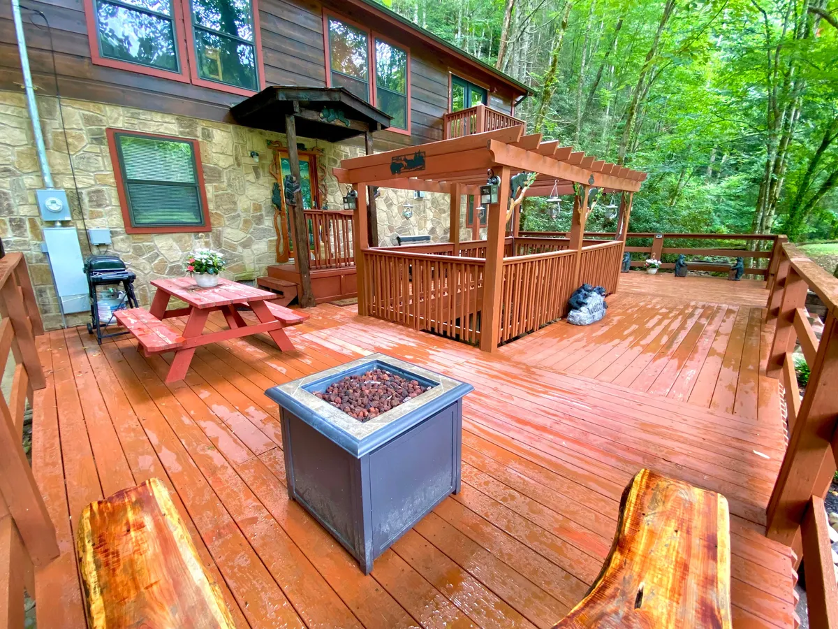 An exterior scene of a cozy wooden deck under a pergola, with a fire pit and picnic table as focal points. The surrounding trees add to the sense of seclusion and connection with nature, while decorative lighting suggests a welcoming space for evening gatherings.