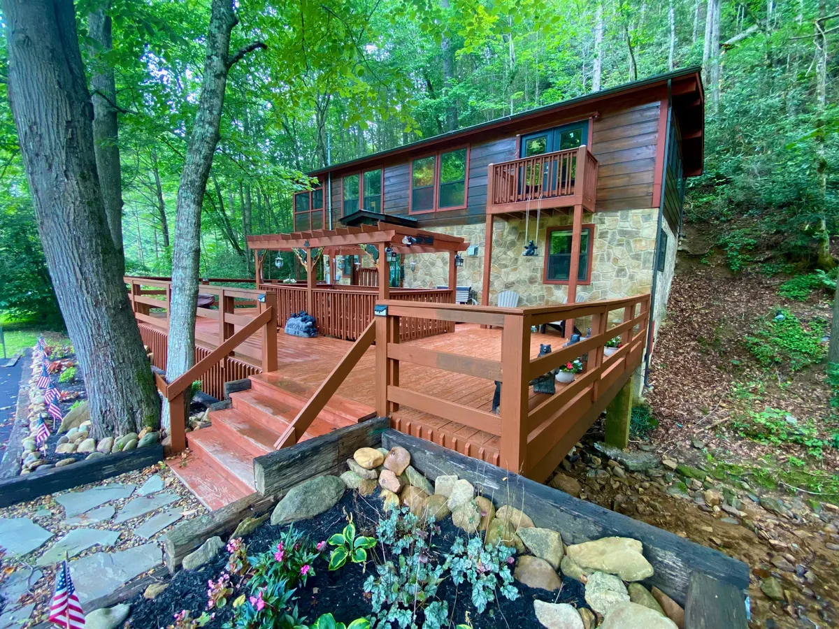 A broad view of the cabin’s exterior showcases a spacious wooden deck, stone accents, flower beds, and mature trees. The inviting outdoor space is perfect for family gatherings, relaxing, or enjoying the scenic natural setting.