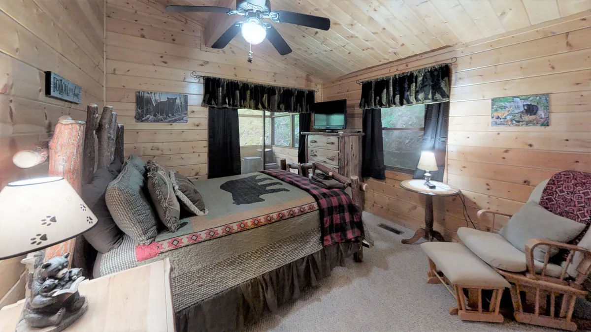 A bedroom with a wooden ceiling, bear-themed bedding, a cozy rocking chair, and a lamp with a bear figurine. The room feels inviting and is filled with natural light, perfect for relaxing or enjoying nature views.